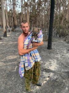 Man caring a koala wrapped in a shirt with burned eucalypt trunks in background