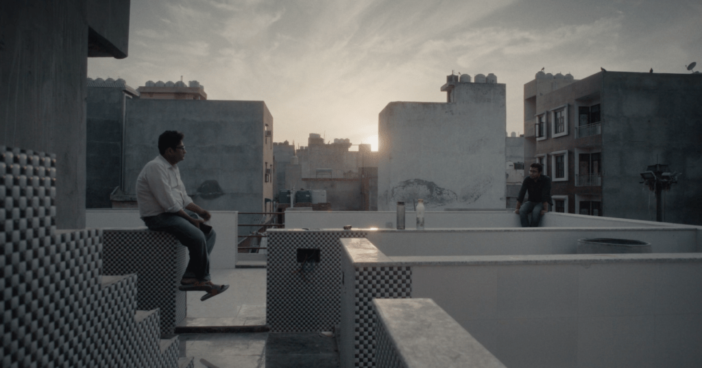 Nadeem Shazad and Mohammad Saud sit facing each other from opposite corners of the roof of a New Delhi apartment building with the sun low on the horizon behind them.