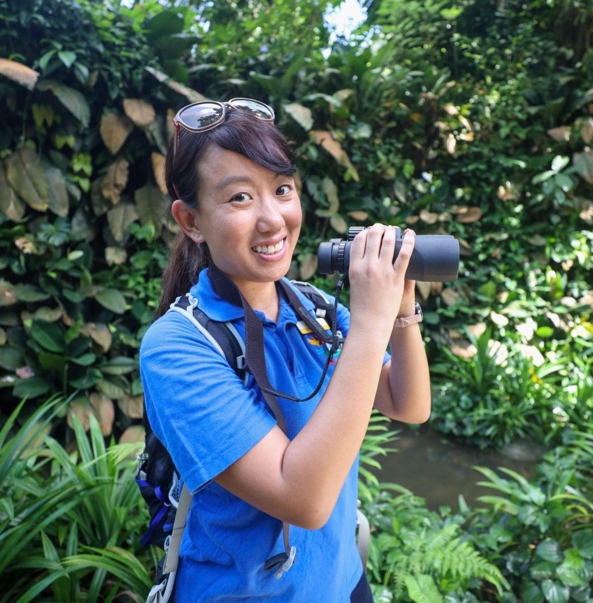 Jessica Lee holding binoculars in a sunny and lush tropical forest